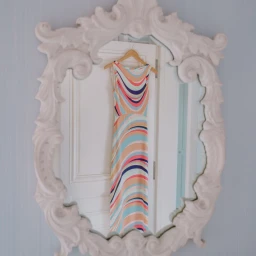 An ornate white framed mirror reflecting a sleeveless multi-colored swirled pattern maxi dress hanging on a wooden hanger against a white door.