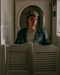 Woman in dark leather jacket and chunky chain necklace behind louvered saloon doors with striped light patterns