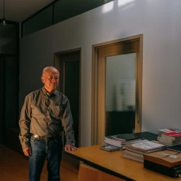An older man standing in an interior office space next to a wooden desk piled with books and magazines, captured in soft directional light.
