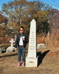 Man wearing red sunglasses and black leather jacket standing beside a marble obelisk in a cemetery.