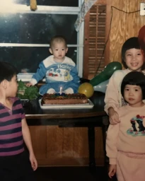 Children gathered around a birthday cake with a single lit candle on a table inside a home