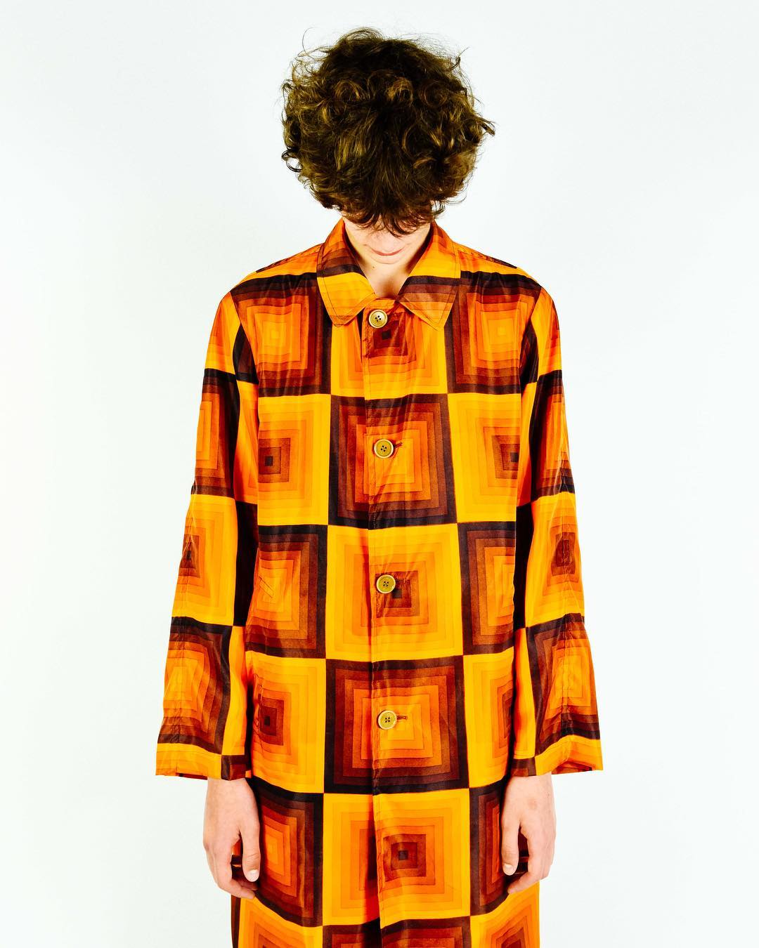 Person wearing a geometrically patterned orange and brown long coat, posing against a white backdrop in a brightly lit setting.