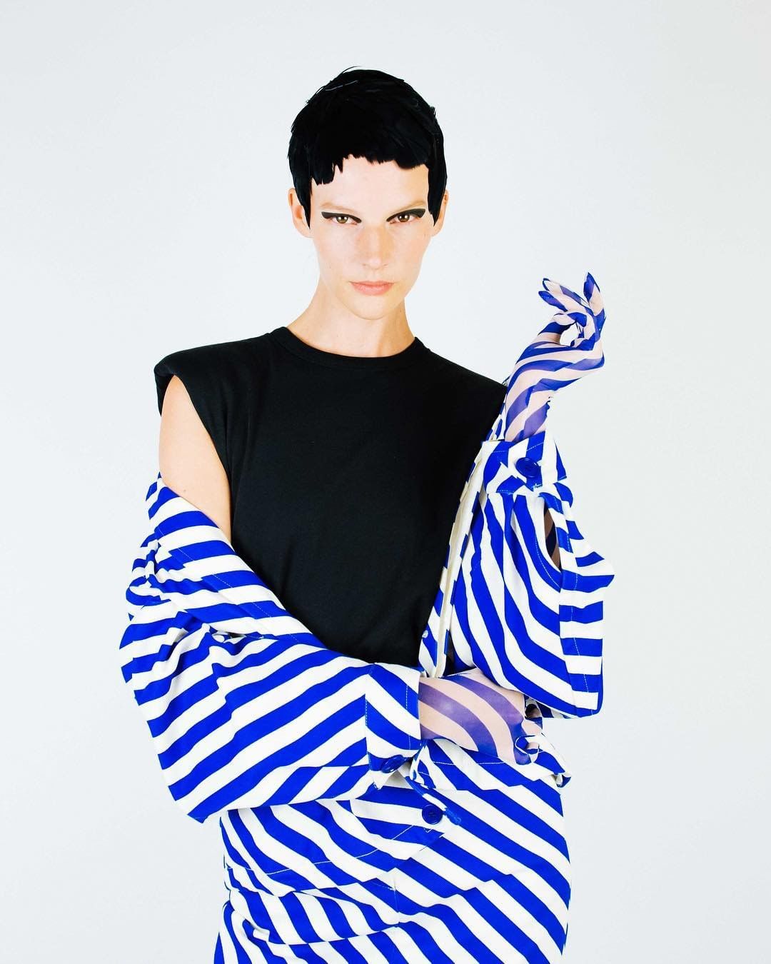 Fashion model poses in a black top and blue-and-white striped jacket and skirt, with matching gloves, against a white backdrop.