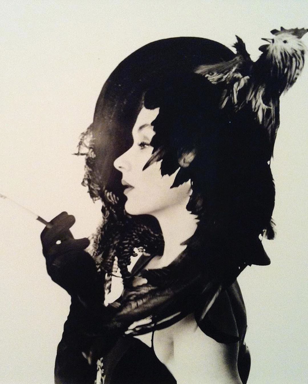 Monochrome portrait of a woman in a feather-trimmed hat with a rooster, holding a cigarette, against a white background.