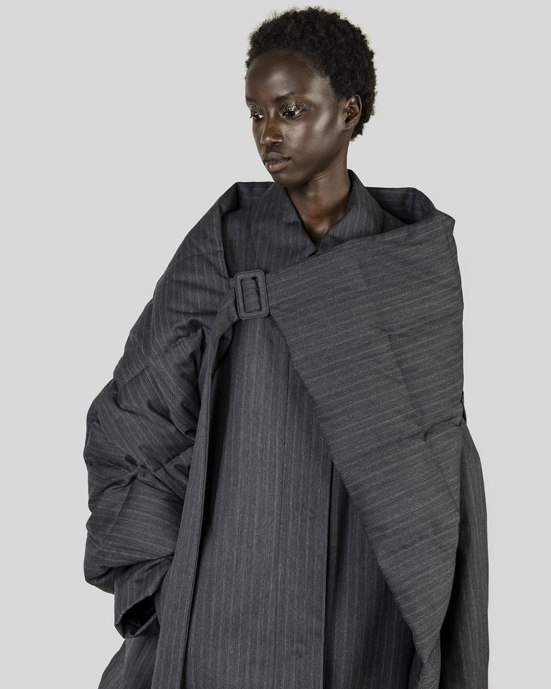 Fashion portrait of a Black woman in an oversized pinstripe coat with a large buckle. The coat's silhouette dominates the frame.