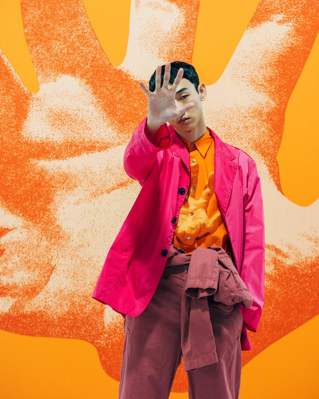 Stylish young Asian man in a pink jacket strikes a pose with an orange patterned background. Fashion-forward and trendy aesthetic.