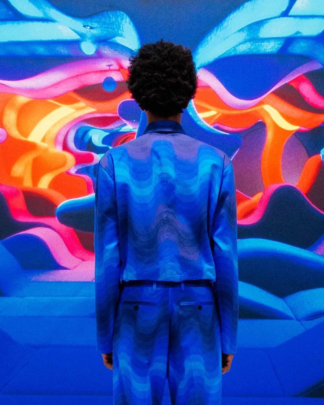 Back view of a person with afro wearing a matching blue patterned suit, standing in front of a blue, orange, and pink abstract painting.