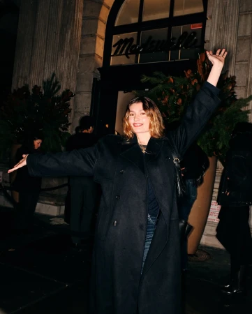 A woman in a dark coat stands in front of a Madewell store at night, with raised arms and other people nearby.