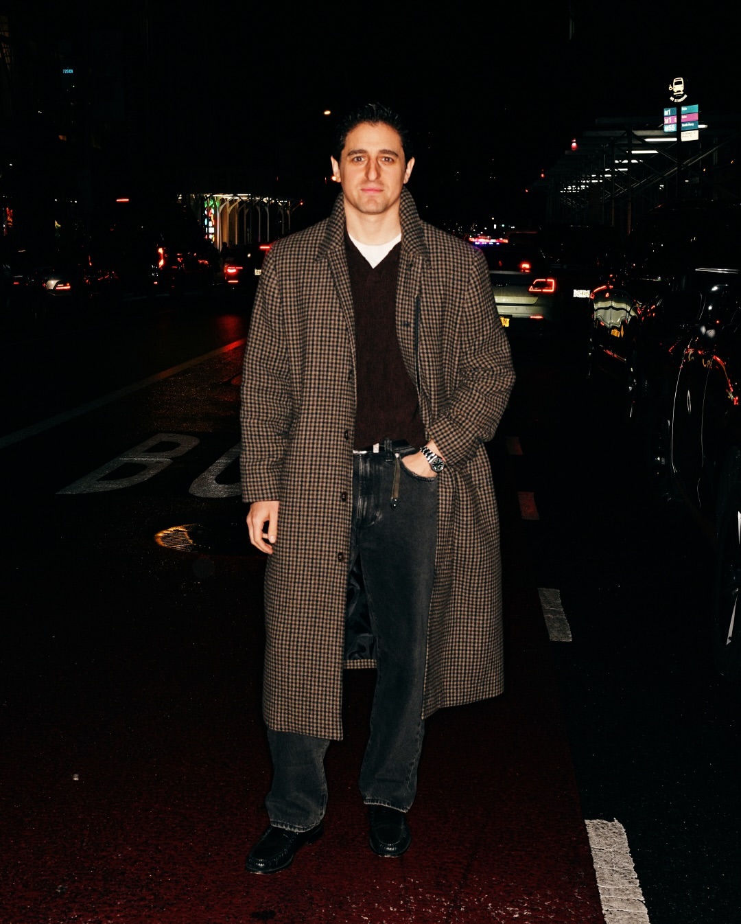 Full-length portrait of an adult male in a patterned coat on a city street at night, with cars and city lights in the dark background.