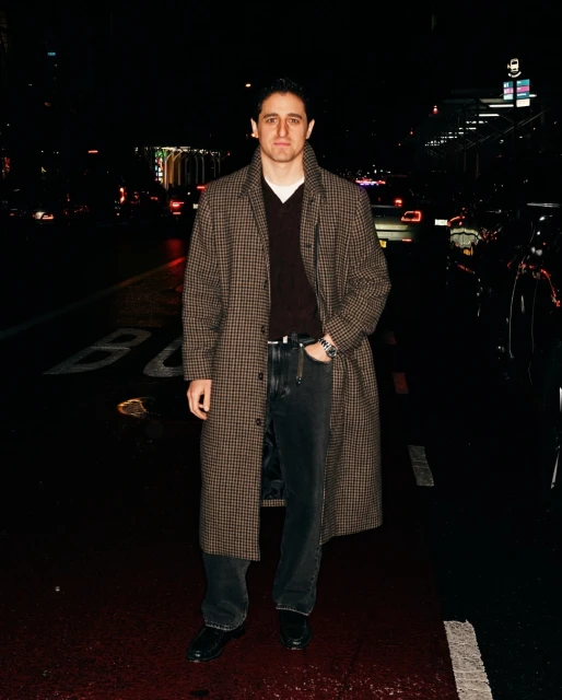 Full-length portrait of an adult male in a patterned coat on a city street at night, with cars and city lights in the dark background.