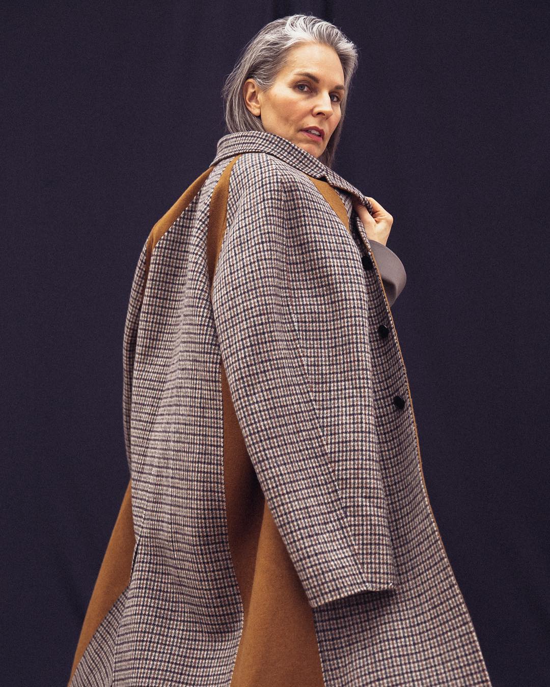 Stylish older woman in a brown and beige plaid coat, posed against a dark backdrop, showcasing a blend of fashion and elegance.