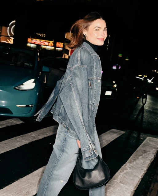 A woman in a denim jacket walks across a zebra crossing at night, carrying a black handbag with cars and buildings in the background.