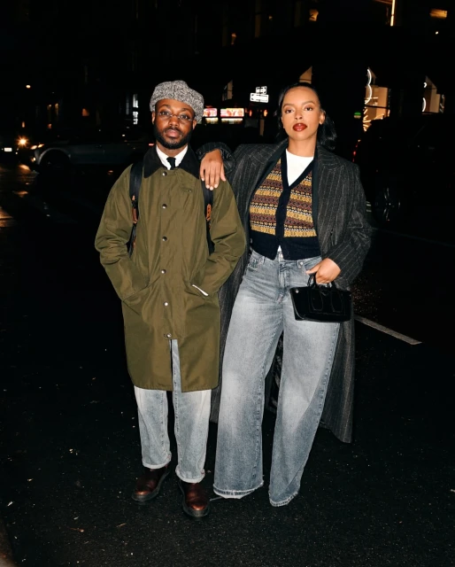 A stylish couple poses at night on a city street. The woman wears a striped coat and the man is in an olive-green jacket and cap.