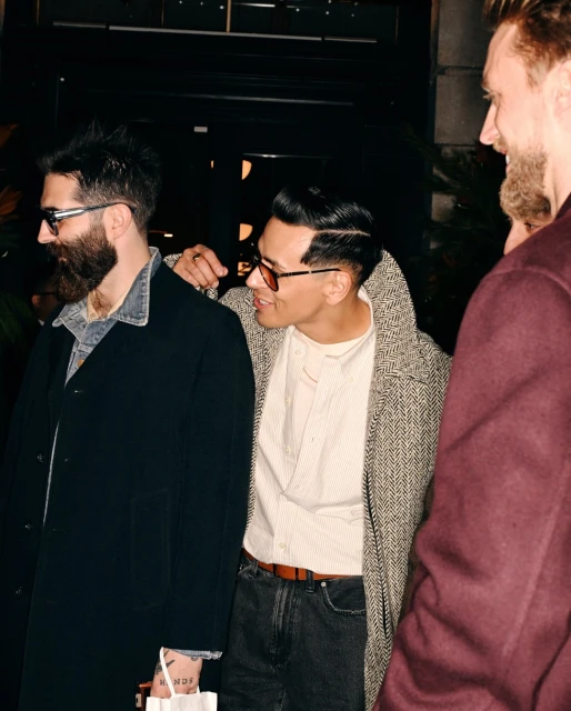 Stylish men in coats and sunglasses stand close together in an urban setting, with one man placing a hand on another's shoulder.