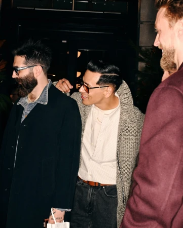 Stylish men in coats and sunglasses stand close together in an urban setting, with one man placing a hand on another's shoulder.