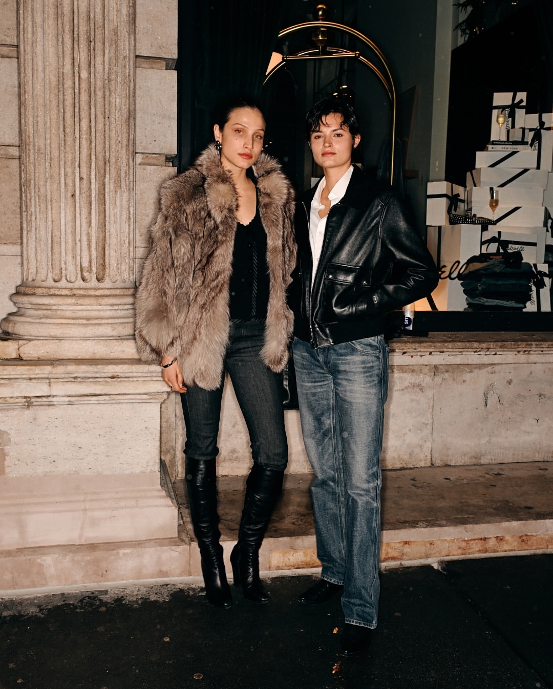Two women in fashionable jackets and jeans pose in an urban setting with a building and a decorative storefront visible in the background.