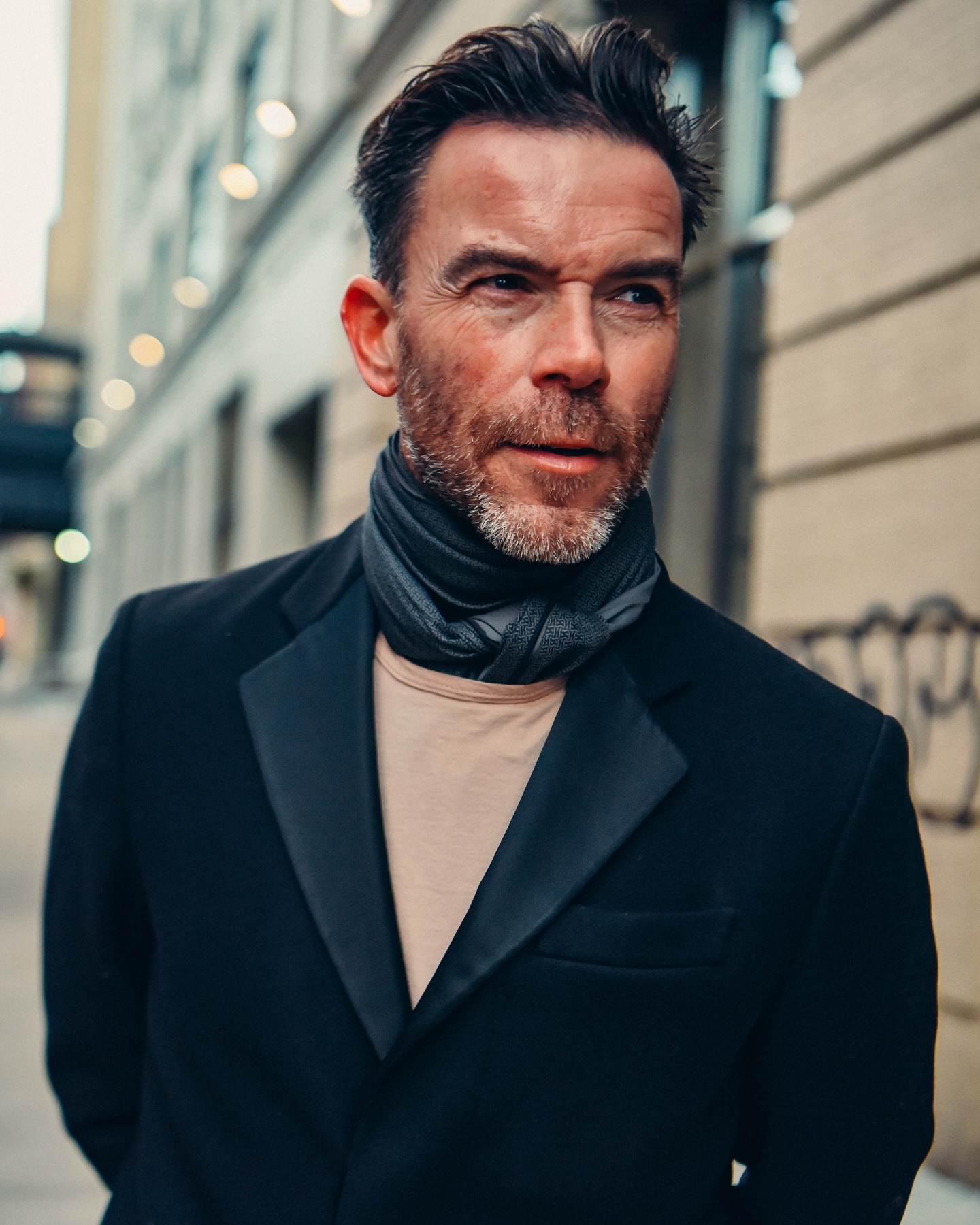 Portrait of a mature man with dark hair and a beard wearing a black coat and grey scarf, standing outside on a city street.
