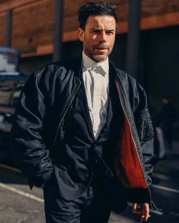 Adult man in a black bomber jacket over a suit and tie walking in an urban setting with brick buildings and city streets in the background.