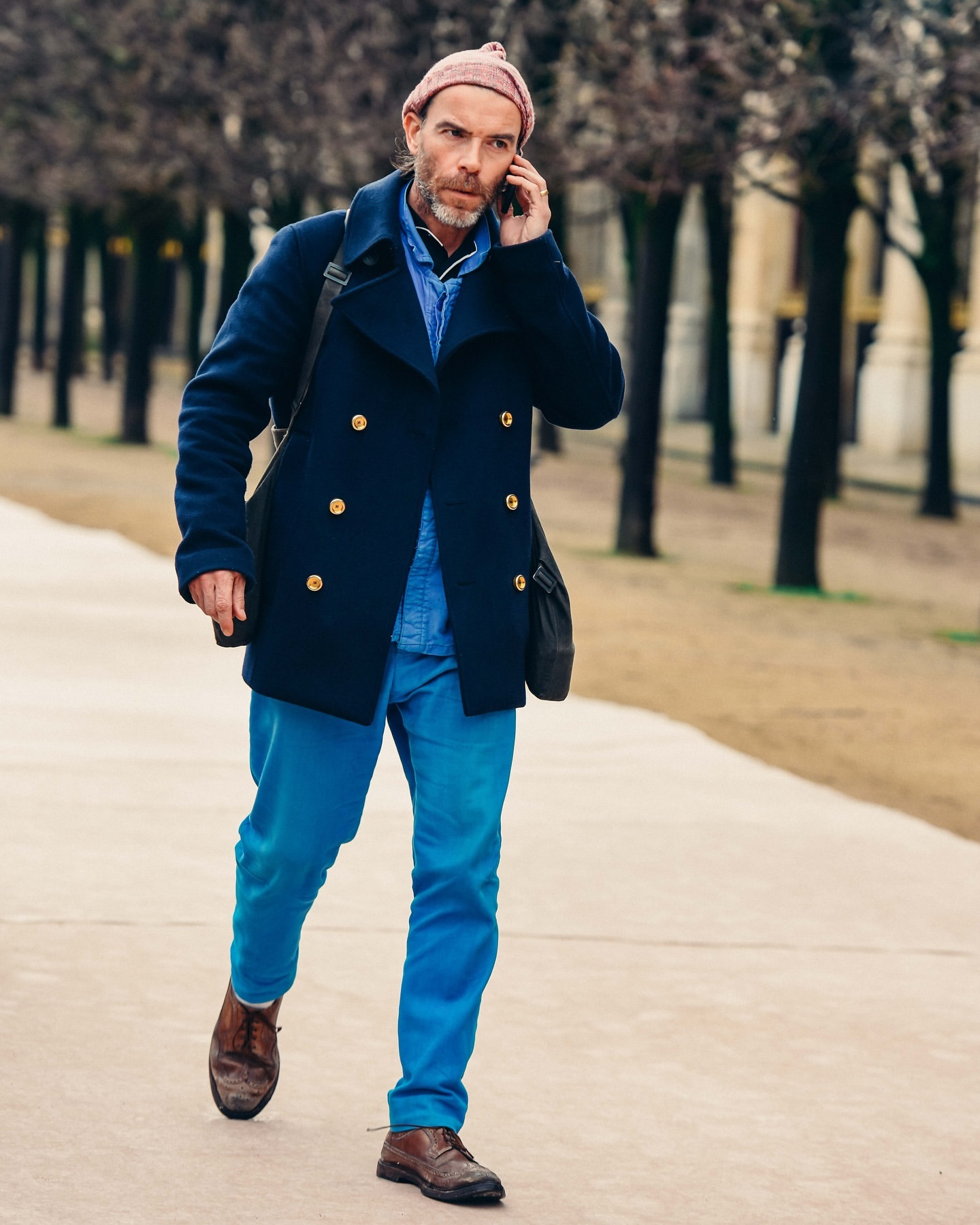 Adult male in navy coat and pink beanie walks on pathway while talking on a smartphone in an outdoor urban setting on a cloudy day.