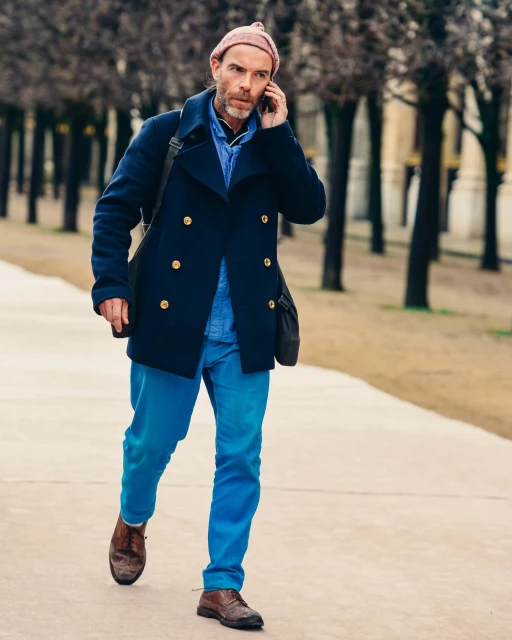 Adult male in navy coat and pink beanie walks on pathway while talking on a smartphone in an outdoor urban setting on a cloudy day.