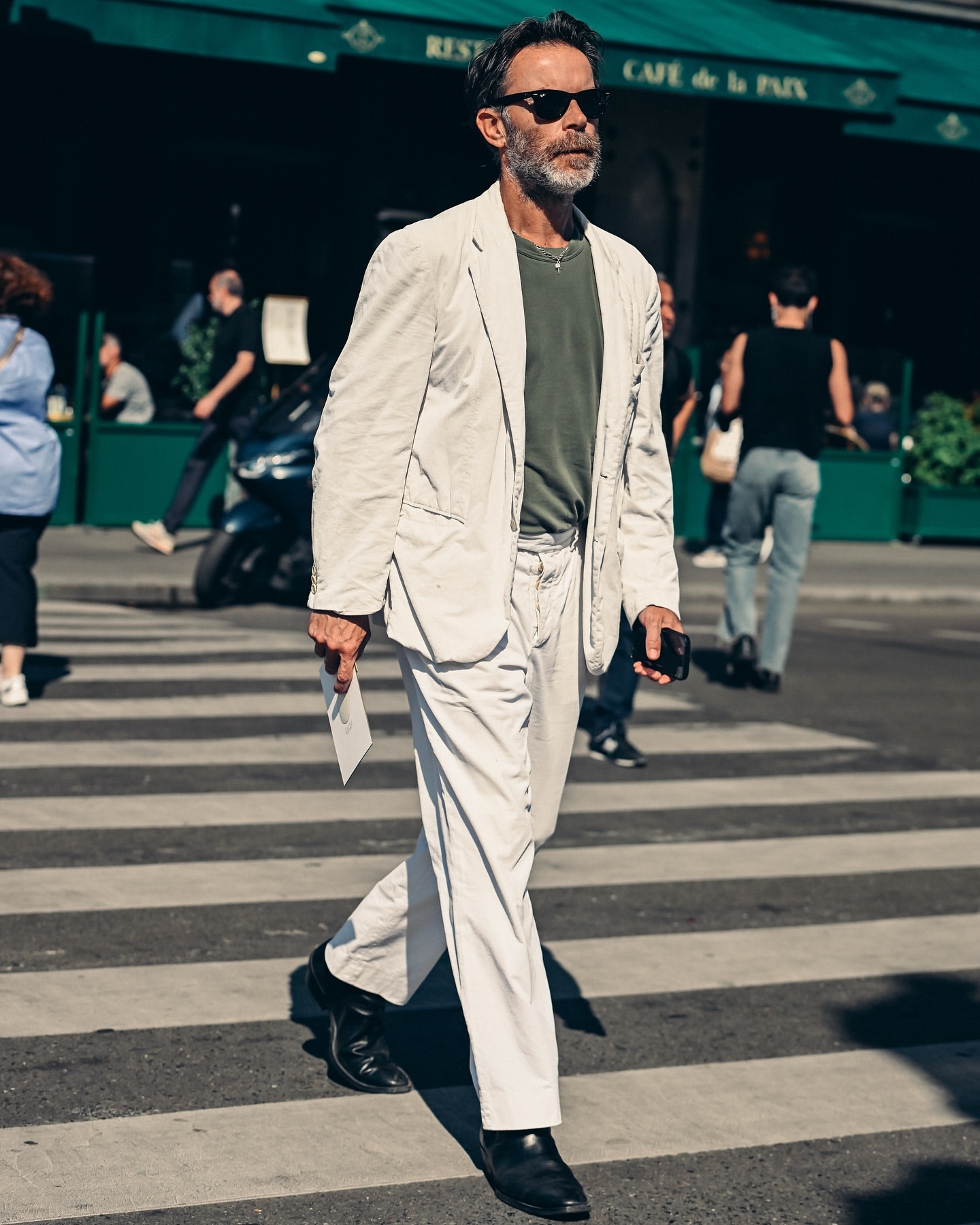 Man in beige suit and sunglasses walks across a zebra crossing in an urban environment, with buildings and cafe in the background.