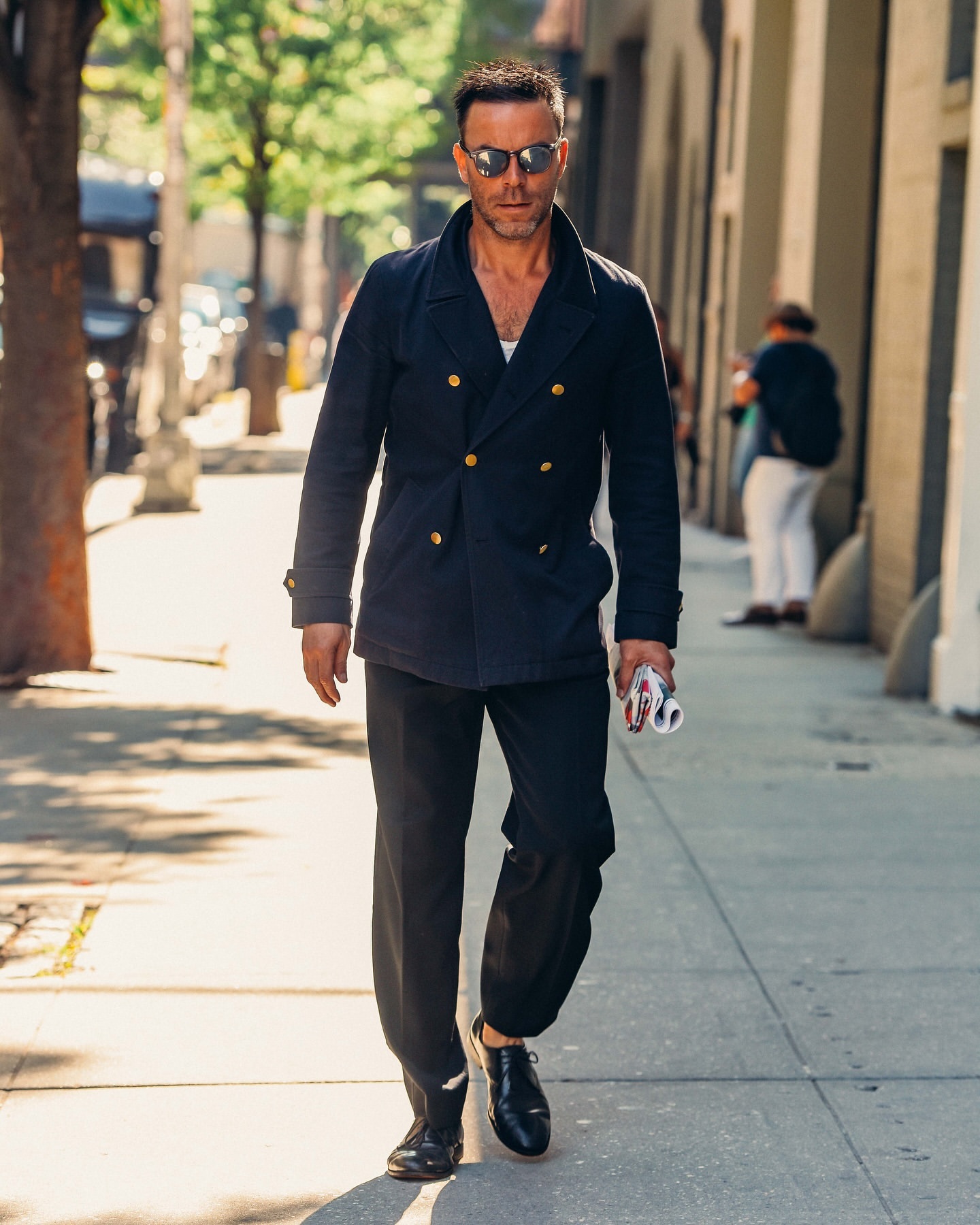 Man in a navy peacoat and black trousers walking on a sidewalk in an urban setting, wearing sunglasses and holding a newspaper.