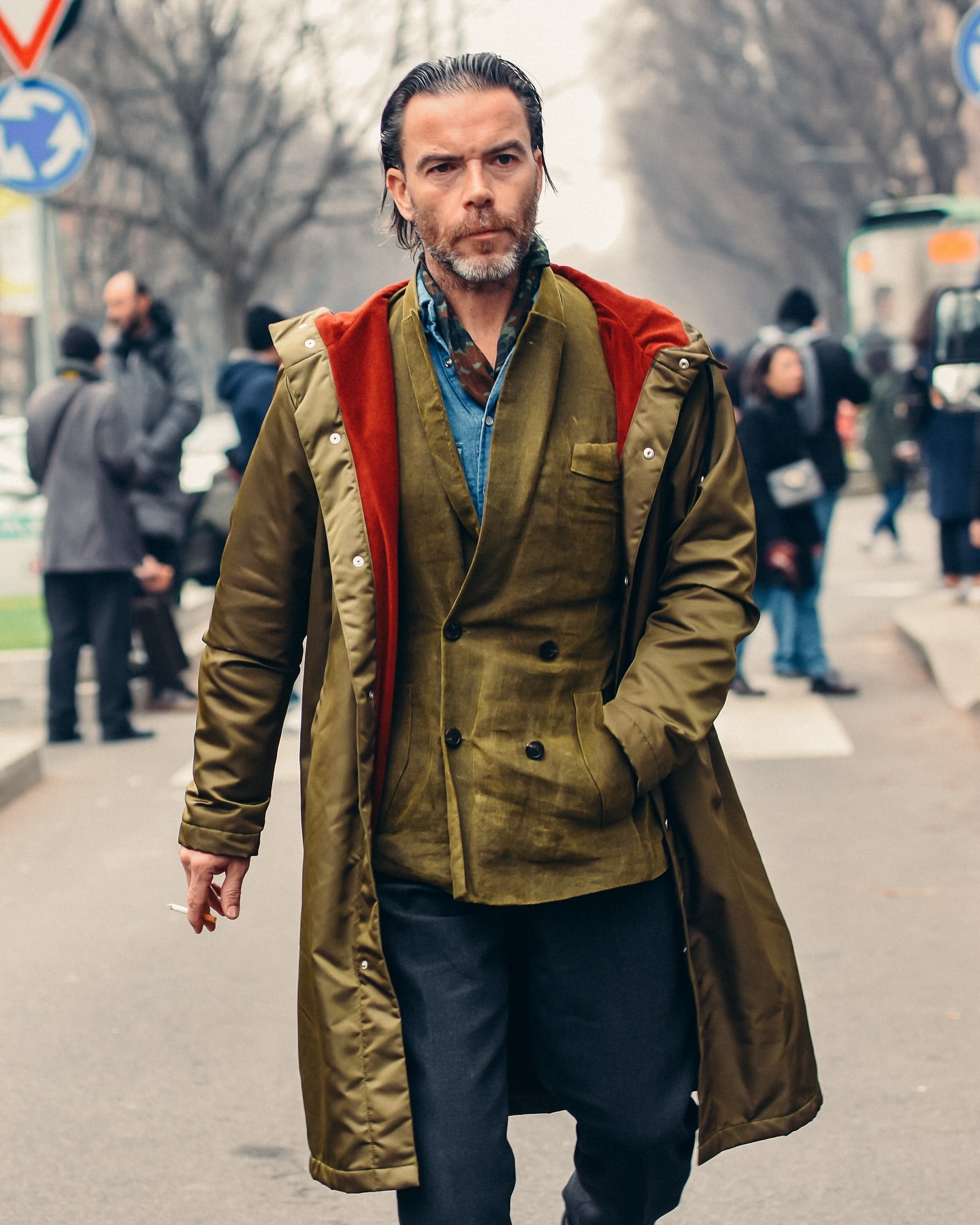 A man in a stylish olive green coat walks on a city street while holding a cigarette, surrounded by blurred pedestrians and buildings.