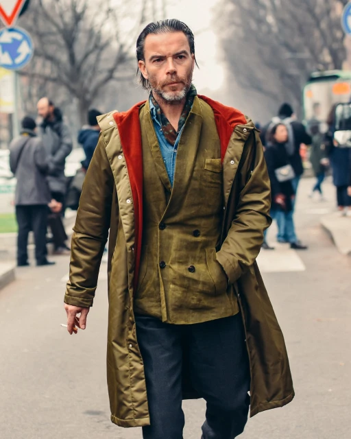 A man in a stylish olive green coat walks on a city street while holding a cigarette, surrounded by blurred pedestrians and buildings.