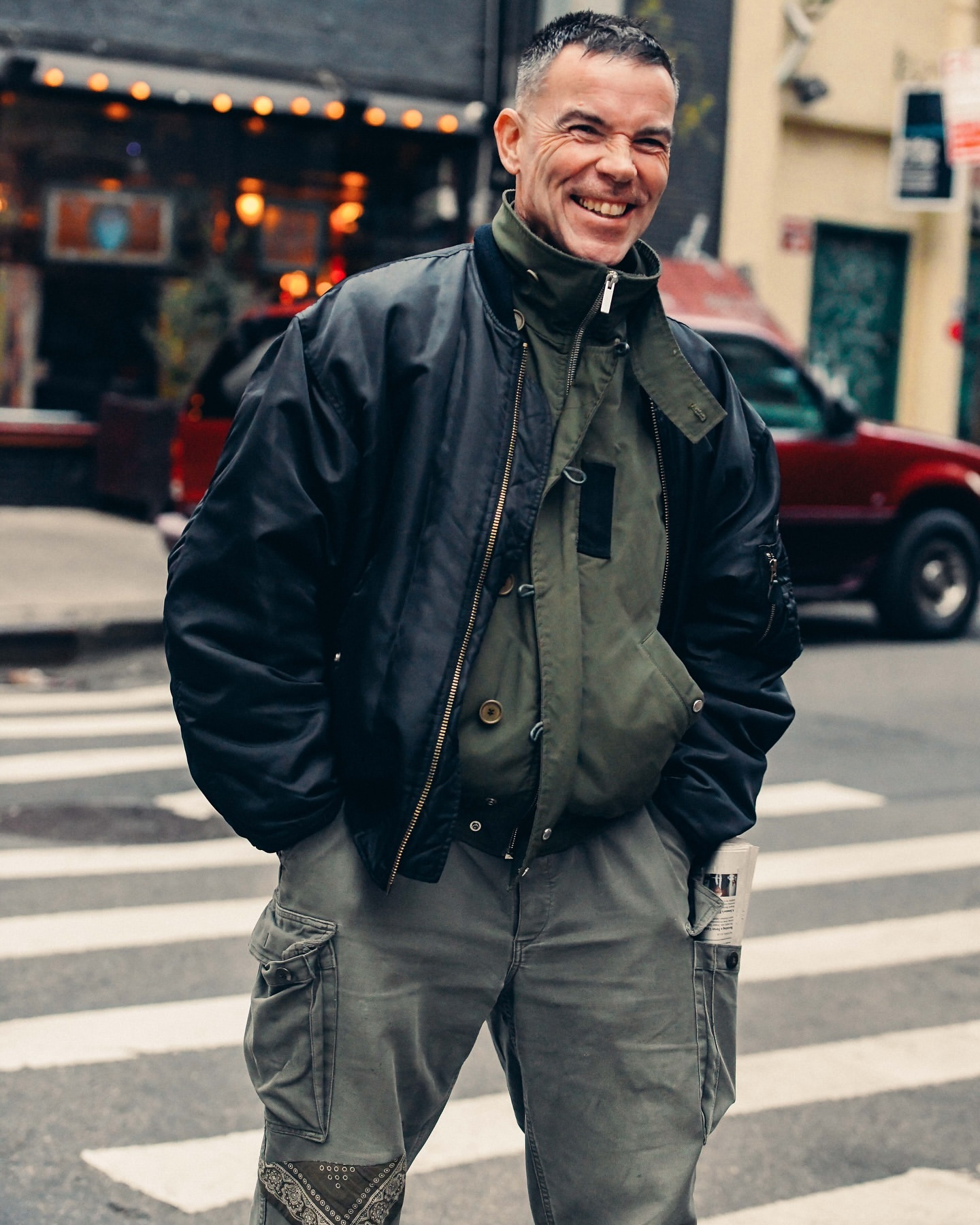 A smiling man with short gray hair in a black bomber jacket and green cargo jacket standing on a city street with a red car behind him.