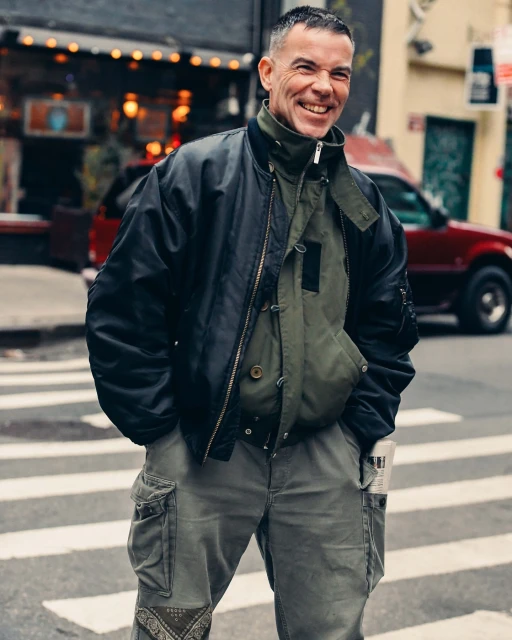 A smiling man with short gray hair in a black bomber jacket and green cargo jacket standing on a city street with a red car behind him.