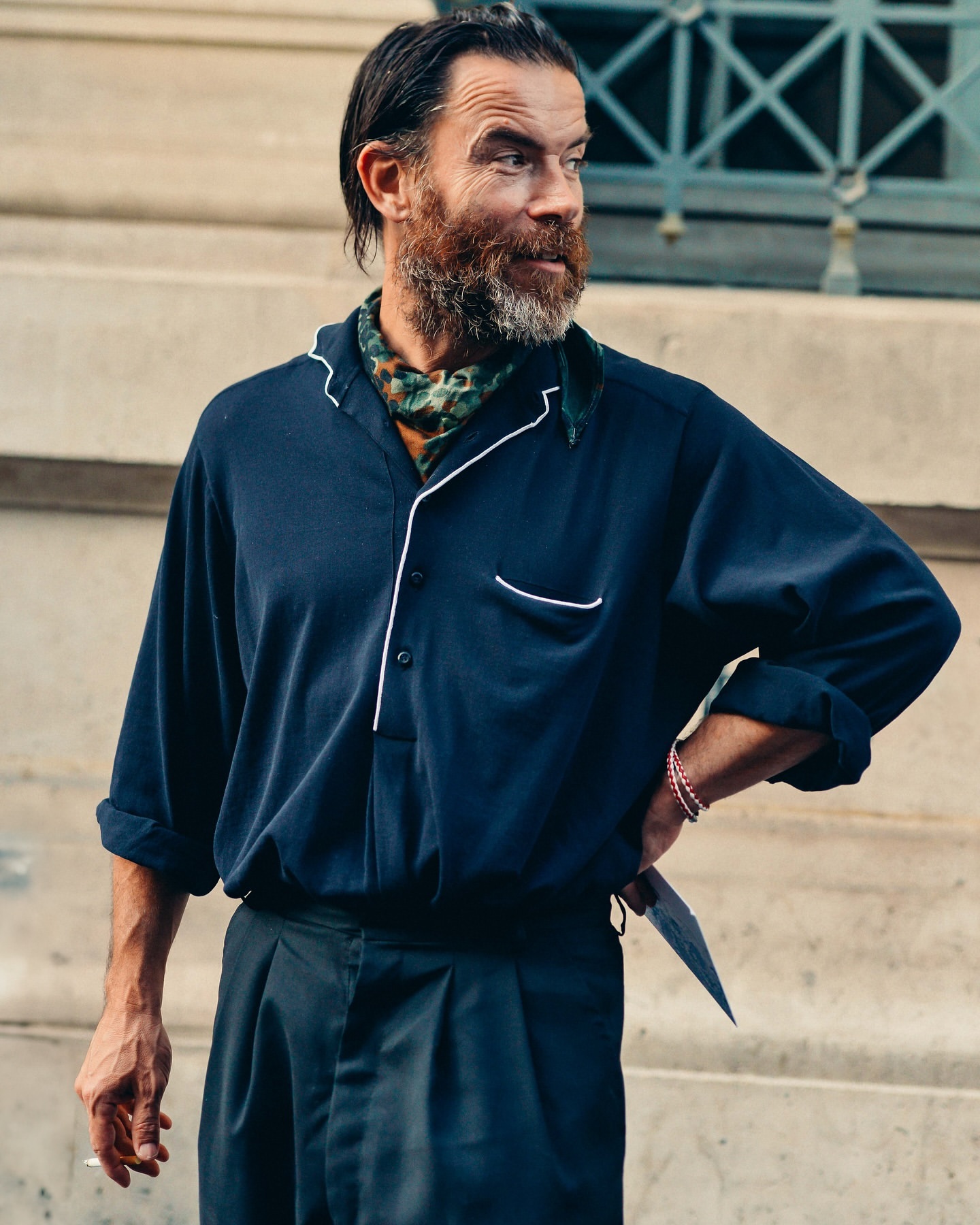 Portrait of an adult male with a beard, wearing a blue collared shirt and dark pants, standing in an urban setting.