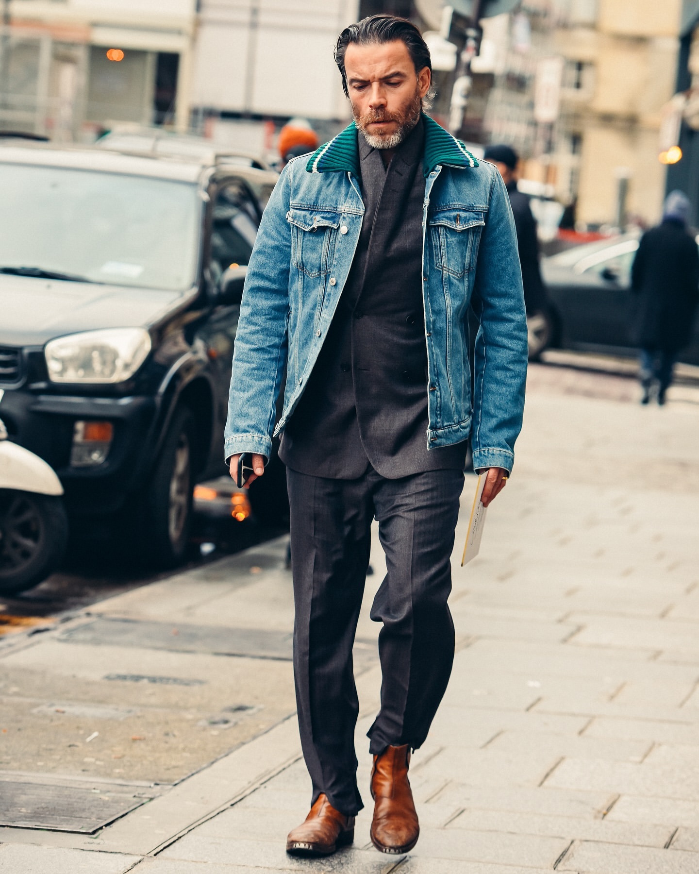 A full shot captures a stylish man in a suit, denim jacket, and brown boots walking in an urban setting, with a black car in the background.