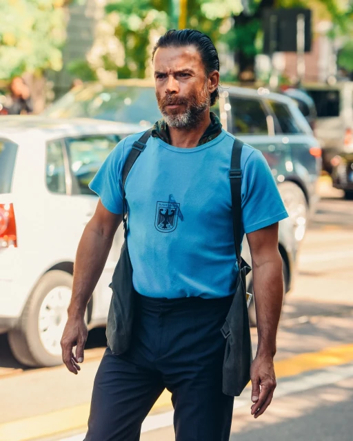 Man in blue shirt walks across a city street on a sunny day with cars and buildings blurred in background. Wearing shoulder bag and dark pants.