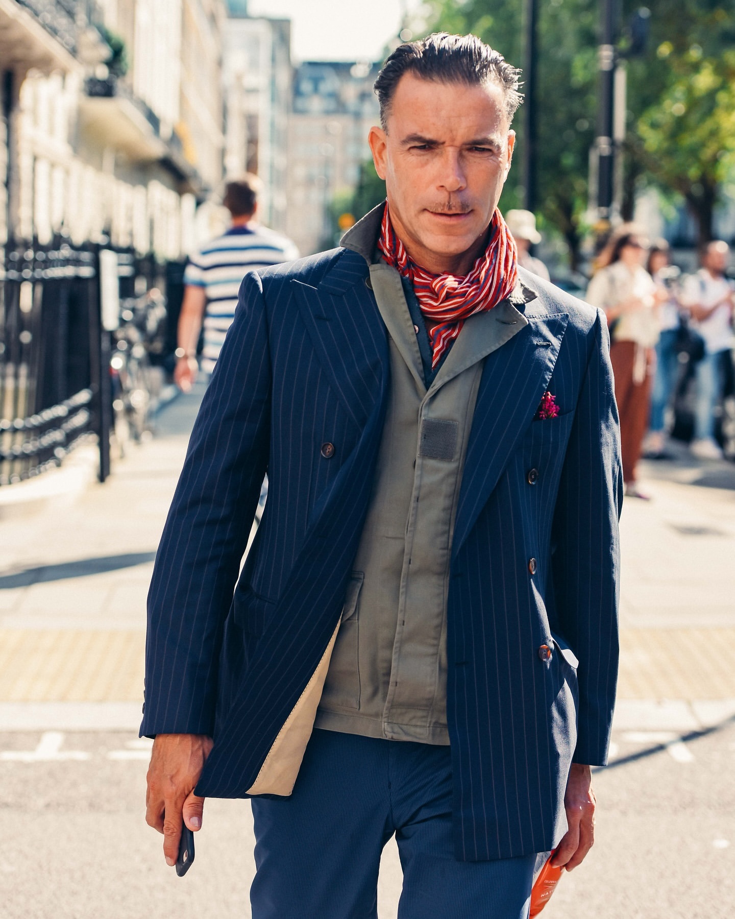 Adult male in pinstripe blazer and red scarf walking on city street, with buildings and pedestrians in background on a sunny day.