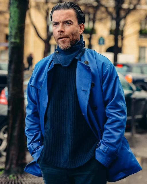 A man in a blue overcoat and a dark sweater stands outdoors in front of a tree with a street and cars visible in the background.