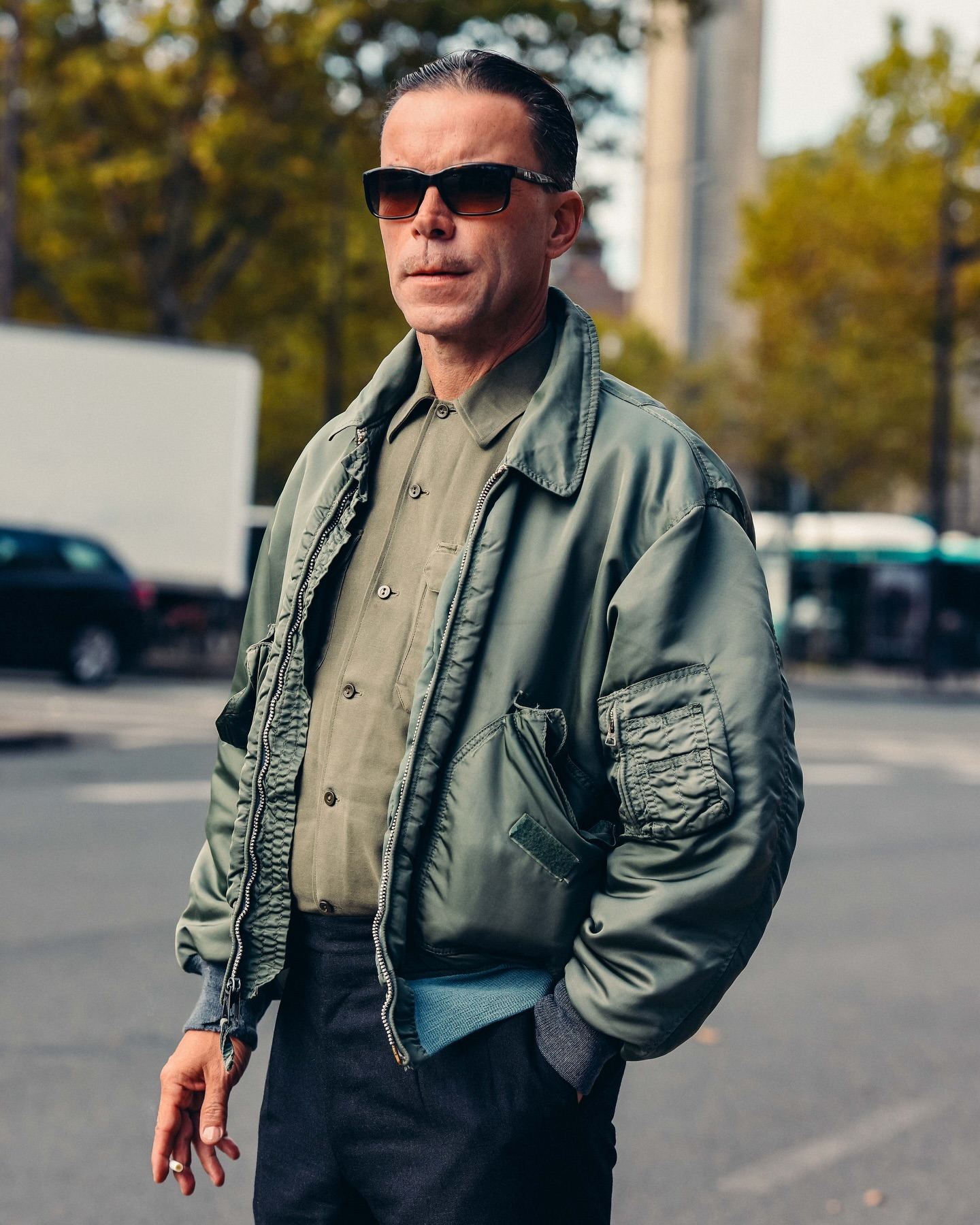 Mature adult man with sunglasses wearing a green bomber jacket poses outdoors on city street with blurred background.