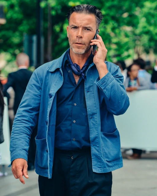 Man in blue jacket on a phone call outdoors. He is wearing a scarf around his neck with a navy shirt and dark pants.