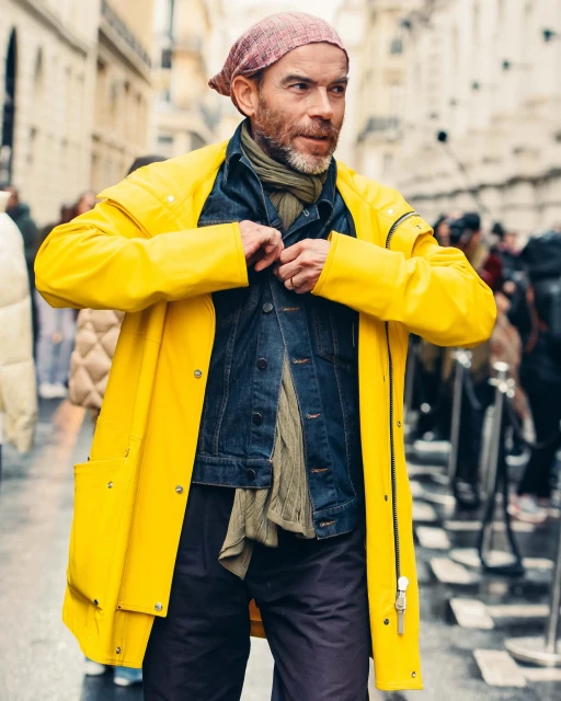 A man in a yellow raincoat and bandana standing on a city street, buttoning his denim jacket, with blurred figures in the background.