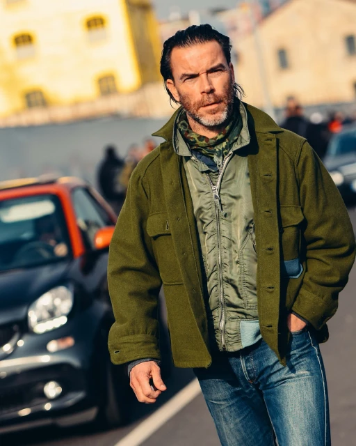 A stylish adult male in an olive jacket walks on an outdoor street with a car in the background, showcasing an urban fashion scene.