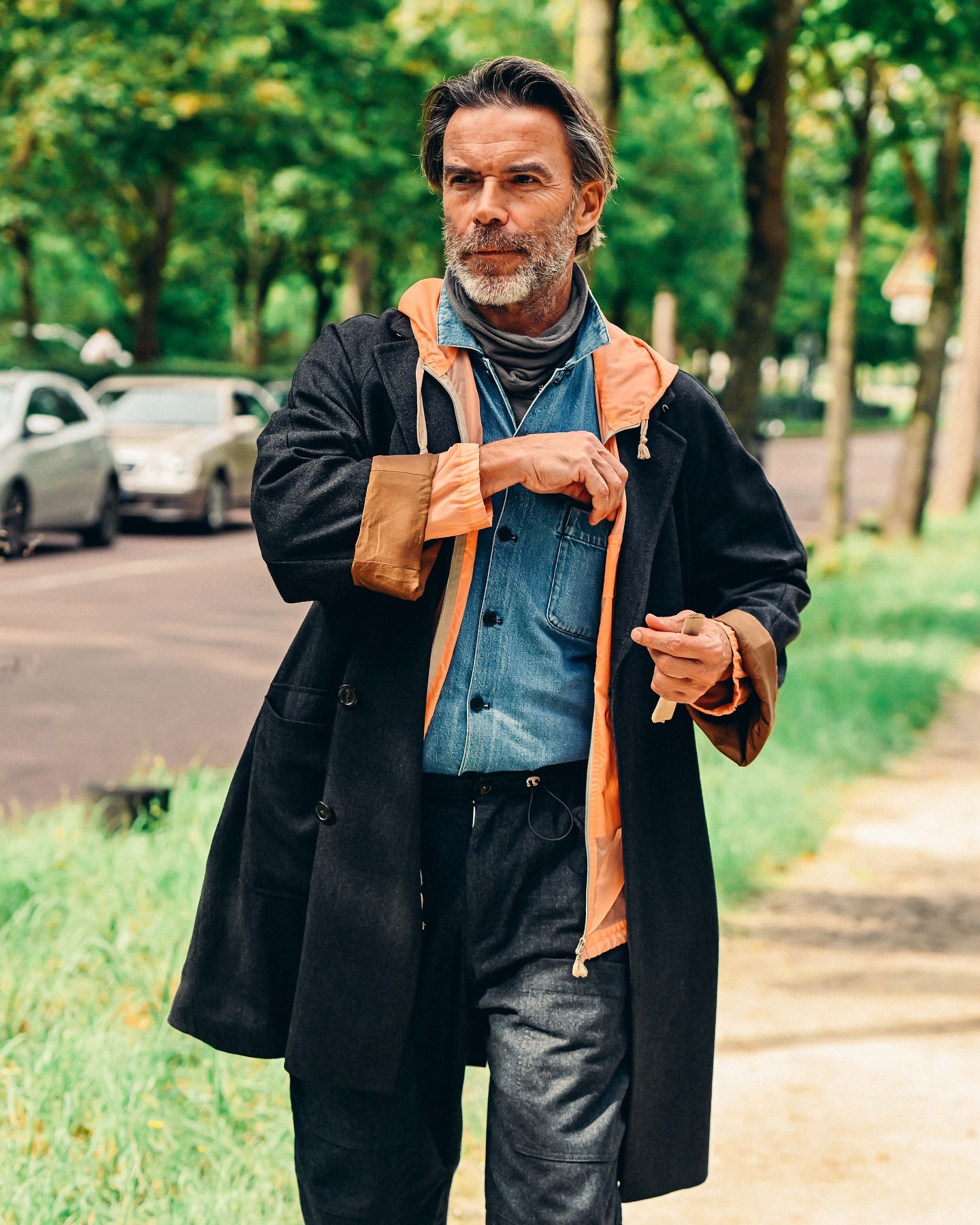 An adult man walking outdoors in a stylish coat, denim shirt, and jeans on a path surrounded by trees and greenery with cars in the background.