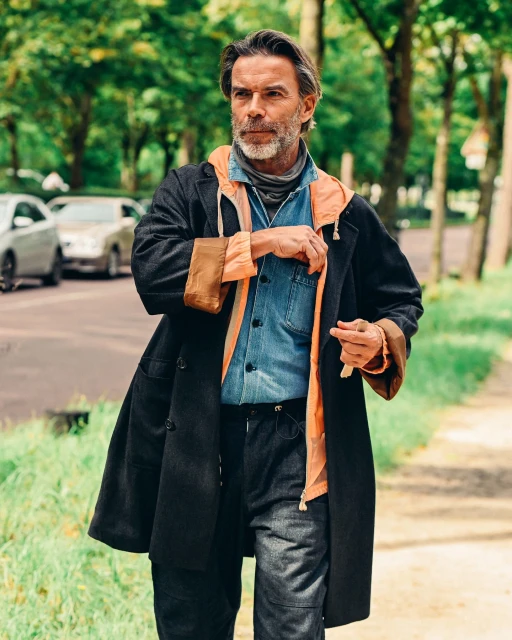 An adult man walking outdoors in a stylish coat, denim shirt, and jeans on a path surrounded by trees and greenery with cars in the background.