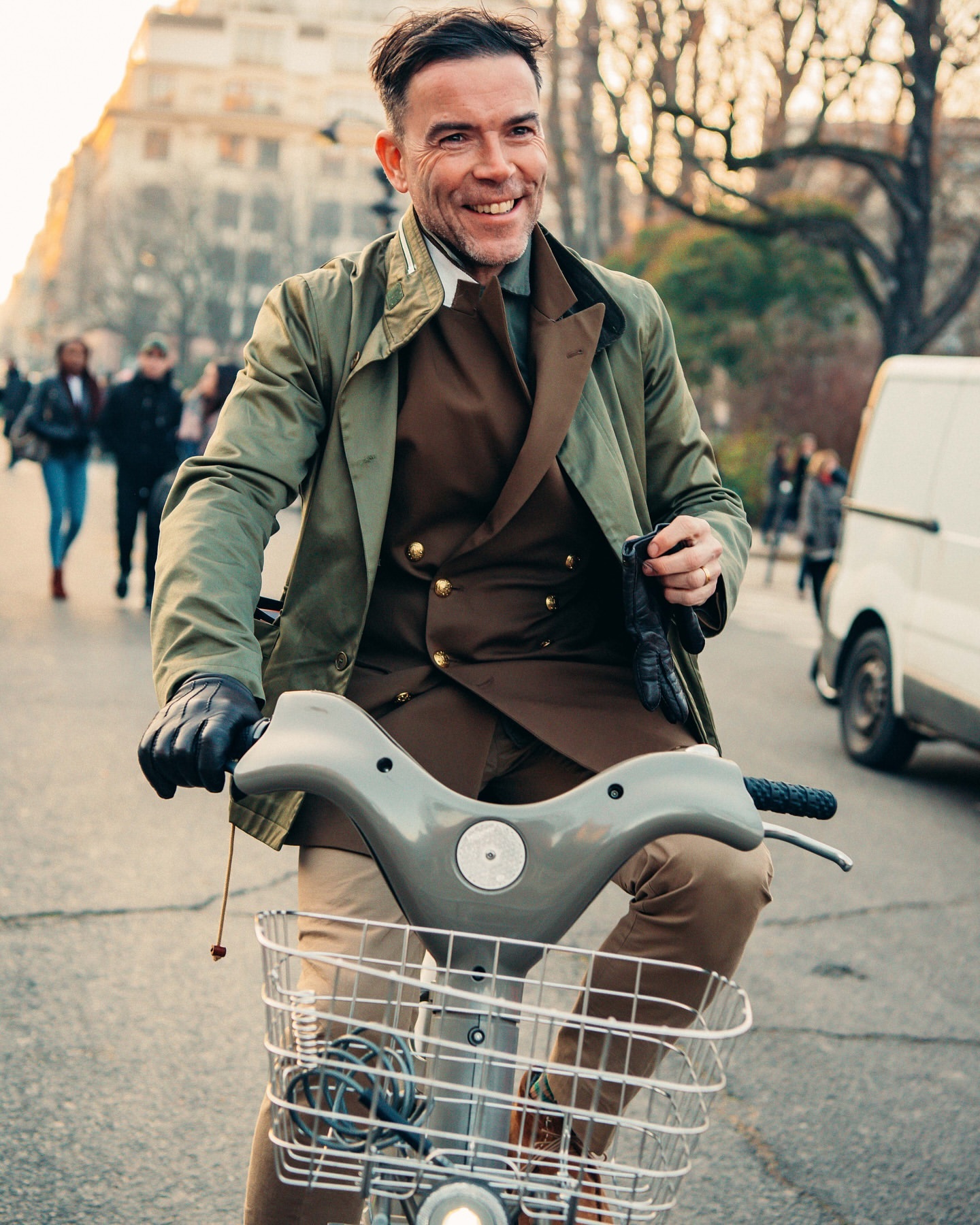 Smiling man rides a vintage bicycle in a European city. He wears a brown suit with an olive coat and gloves; buildings line the street.