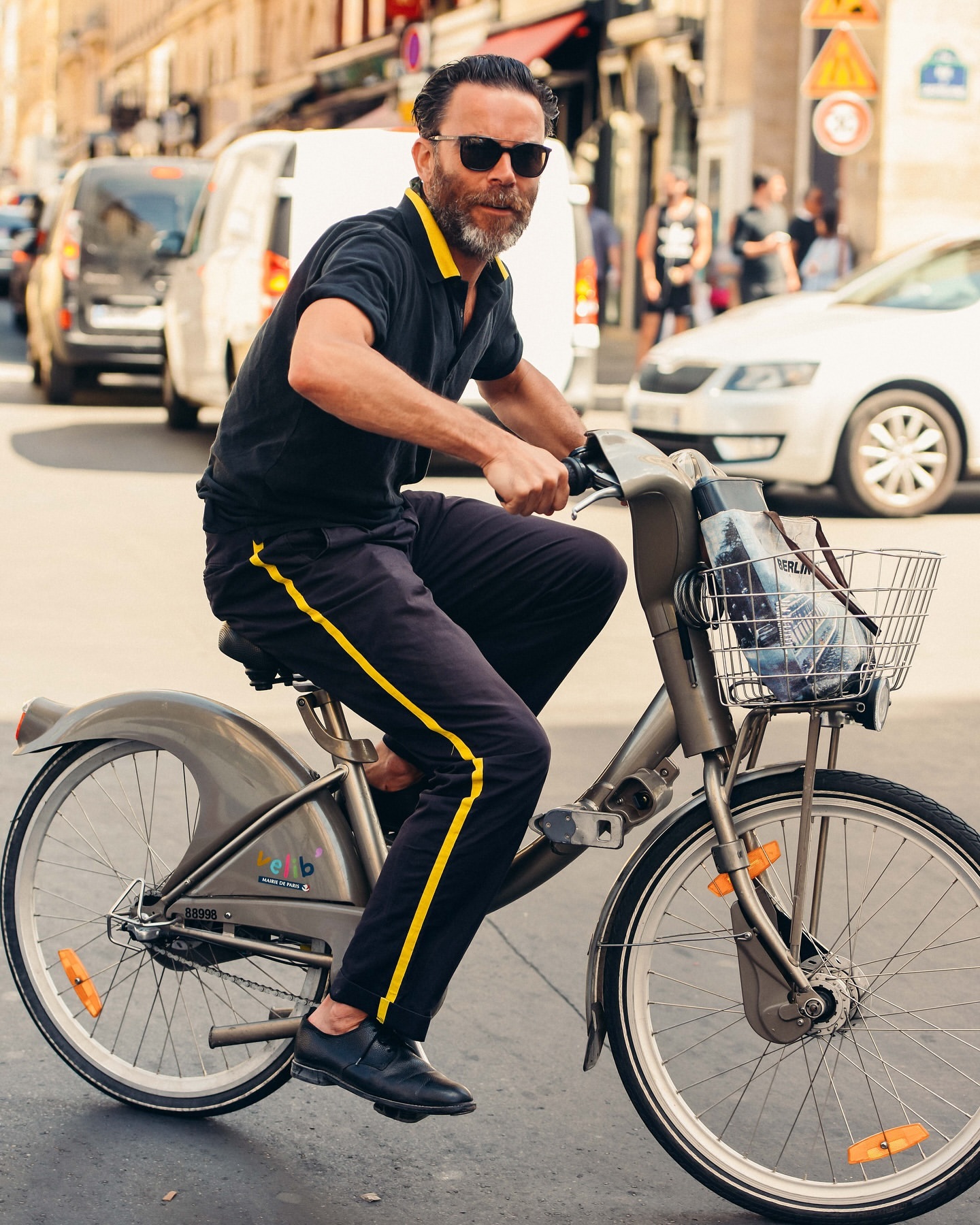 Adult male rides a city bicycle on a sunny street in Paris. He wears a black polo shirt, dark pants, and sunglasses, with cars behind him.