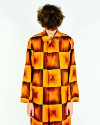 Model with curly brown hair wearing a long buttoned coat with an orange and brown geometric square pattern on a white background