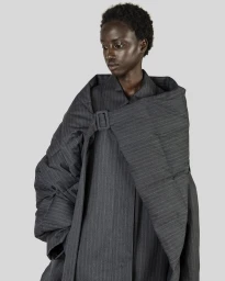Black woman wearing a dark grey pinstripe coat with a buckled wrap collar against a plain grey background