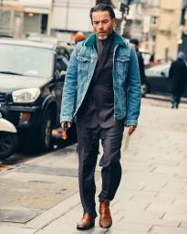Man walking on a city street wearing a denim jacket over a dark suit and brown leather boots