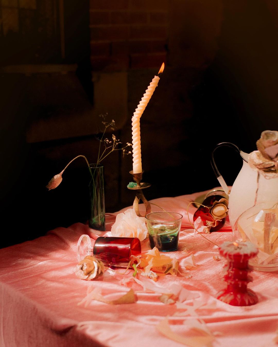 Still life with a twisted candle in a brass holder, glass vases, and rose petals scattered on a pink tablecloth against a dark background.