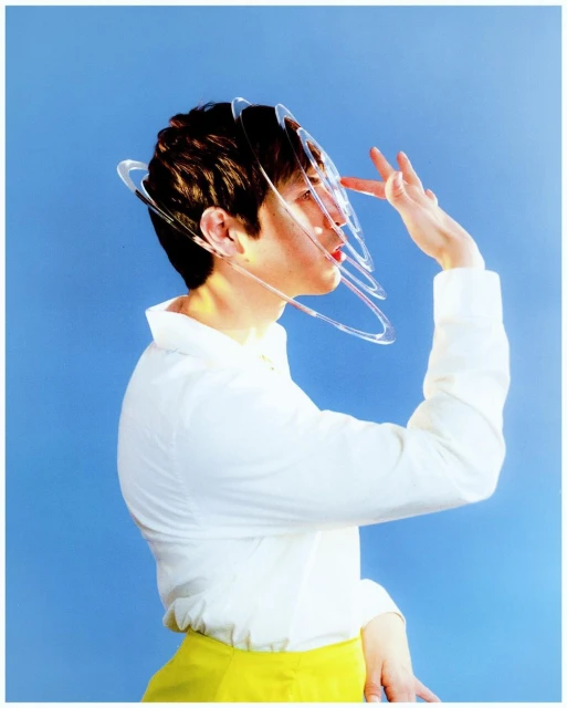 Young adult poses in profile with futuristic clear headgear. White shirt and yellow skirt against a bright blue background.