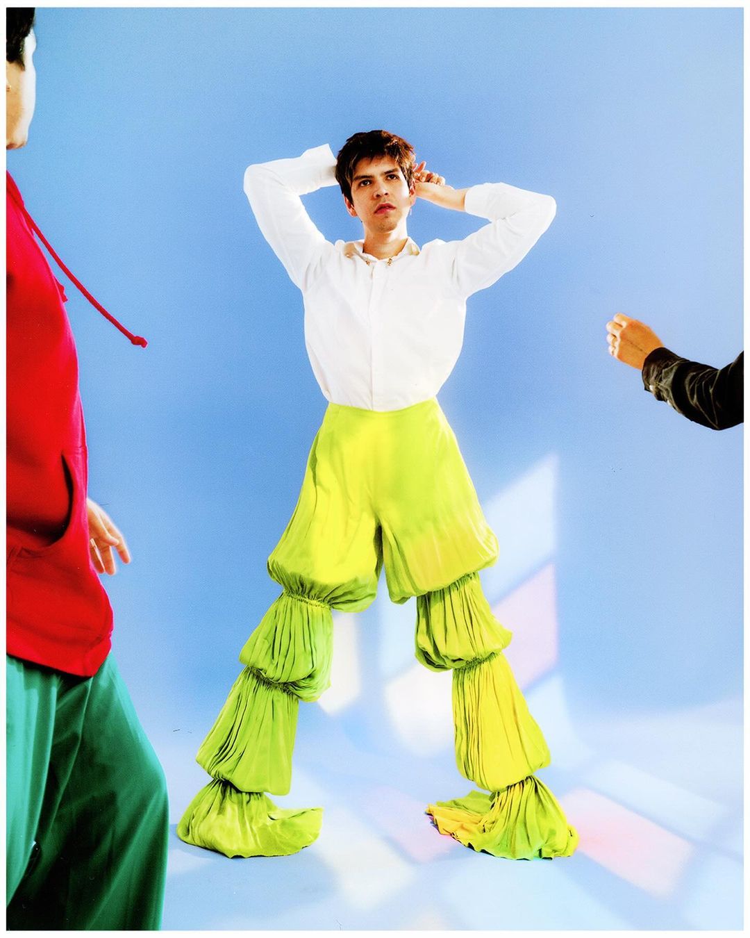 Stylish adult male poses in a photo studio with bright light. He wears ruffled chartreuse tiered pants, and two people stand on either side.