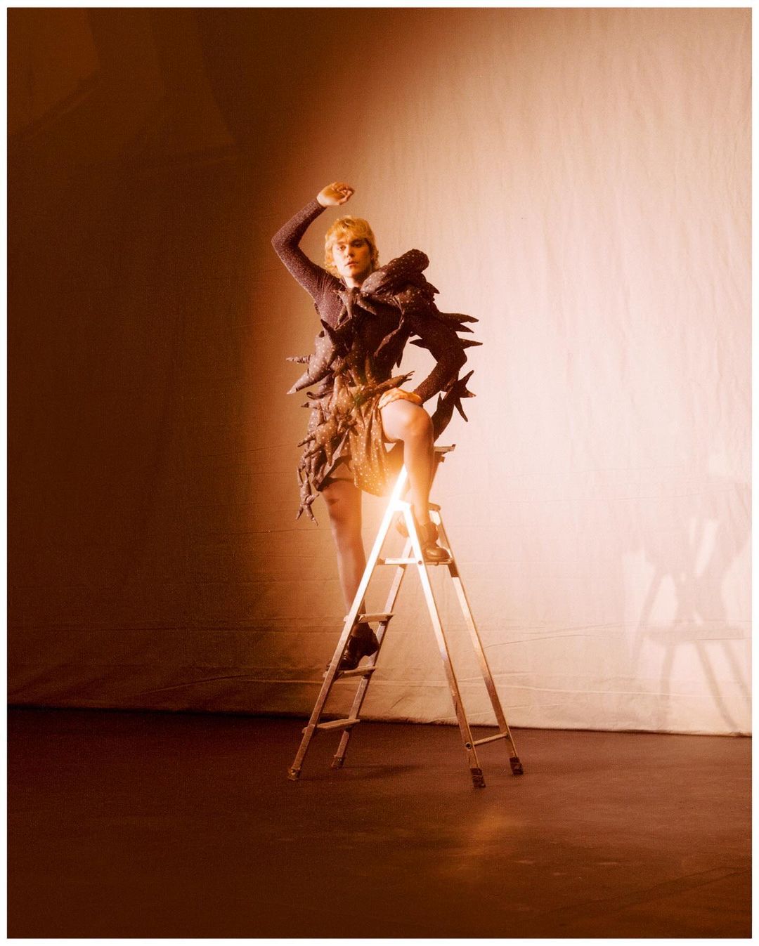 Fashion photo of a blonde adult female model in an avant-garde dress, climbing a ladder in a brightly lit photo studio setting.